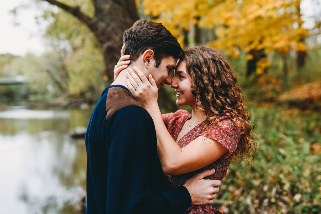 couple touch foreheads in front of fall colors on White River in Noblesville during their Potters Bridge Engagement session