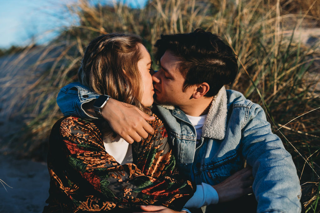 young blonde woman kisses young dark haired man in front of dune grass during their Indiana Dunes engagement photos
