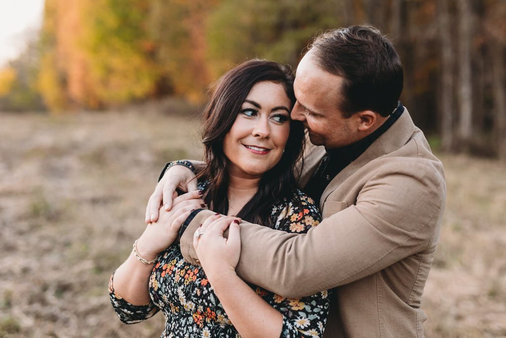 Man in brown suit jacket hugs woman in floral dress in field during their starkey park engagement photos