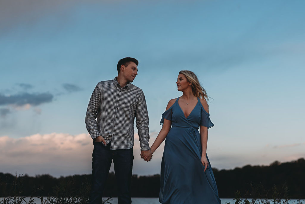 blonde woman in blue dress holds hands with man in gray shirt during blue hour at their eagle creek photo session