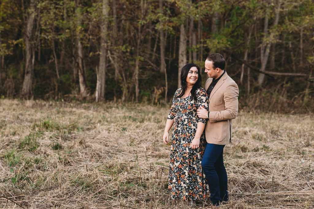 man holds woman lovingly from behind in field in front of forest during their starkey park engagement photos