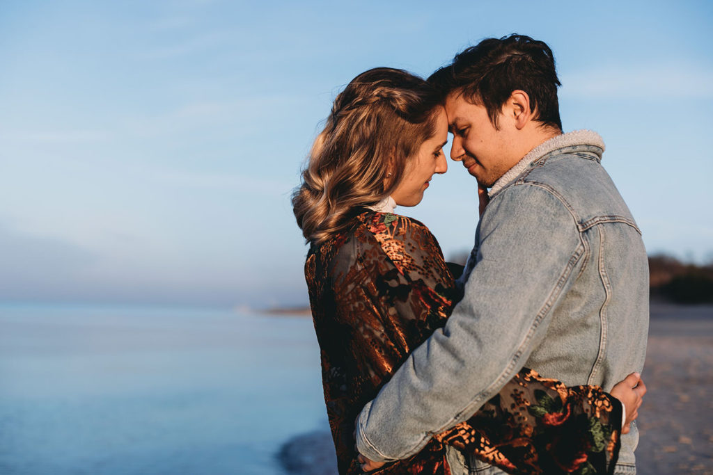 young couple cuddle and touch foreheads together next to Lake Michigan during their Indiana Dunes engagement photos
