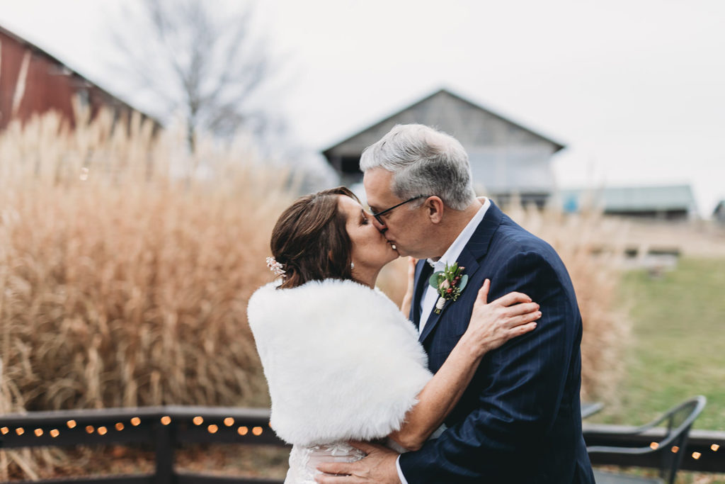 bride and groom kiss on chilly day in front of grass during their traders point creamery elopement