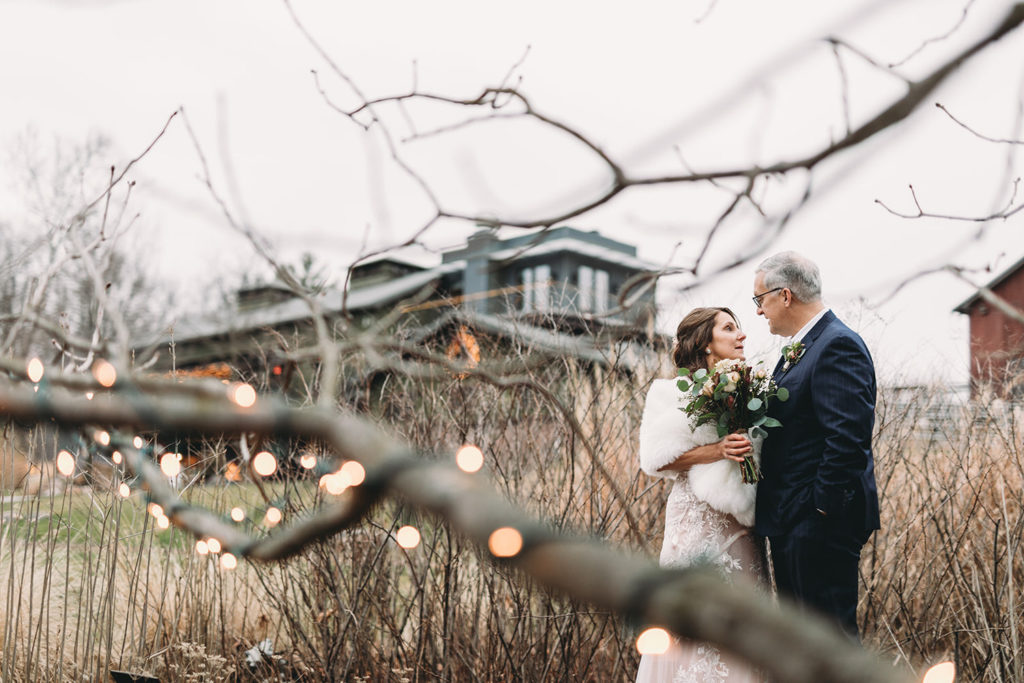 framed by tree branches and standing in front of tall grass and a rustic building a bride and groom hug during their traders point creamery elopement