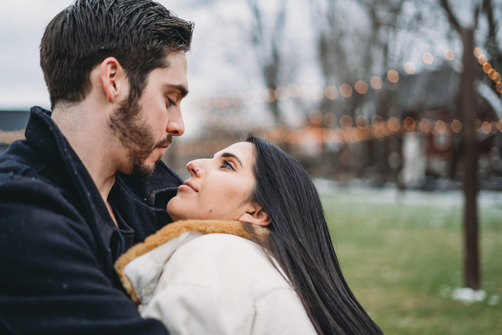 man dips woman in front of bokeh while wearing winter coats during their mustard seed gardens engagement photos