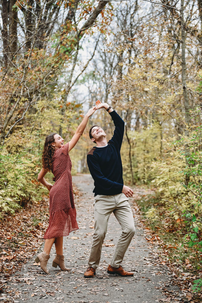 woman spins man on trails surrounded by fallen leaves during their Potters Bridge Engagement session
