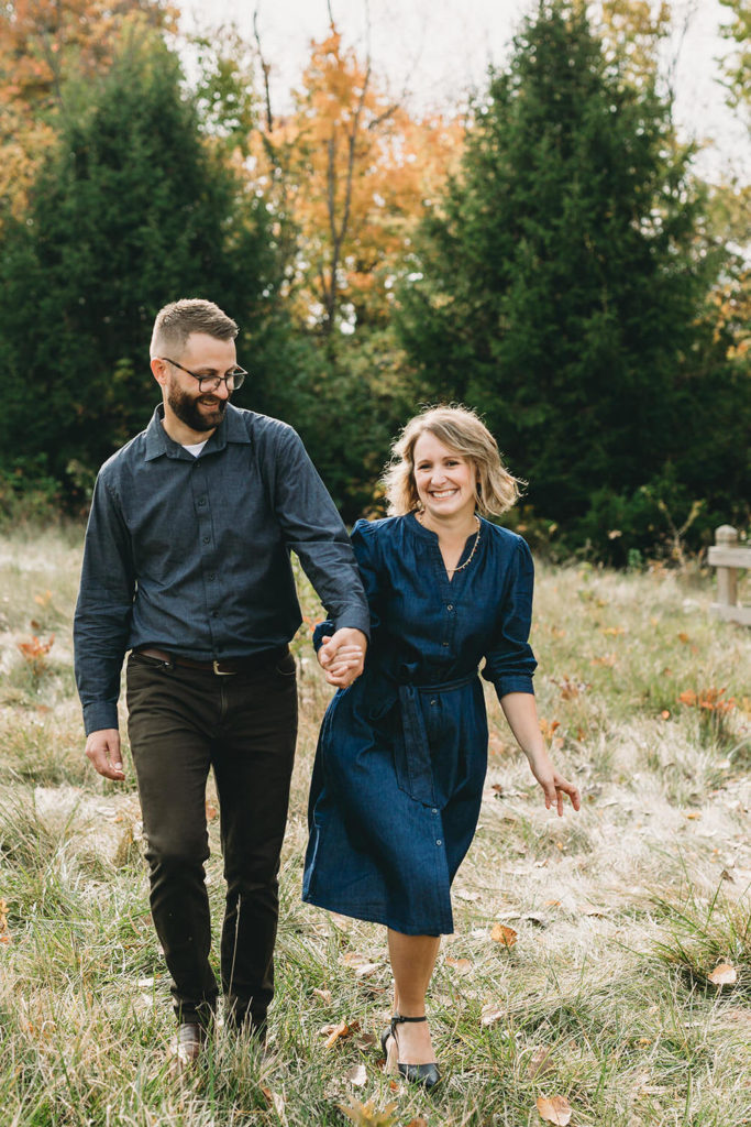 husband and wife hold hands and walk through field captured by a Carmel Family Photographer