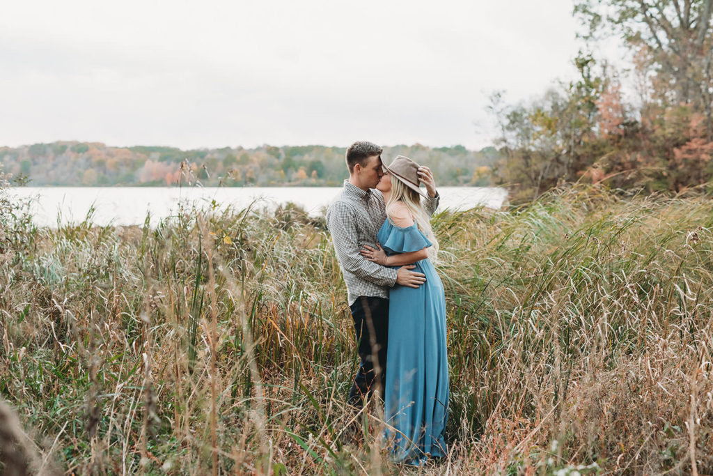 blonde woman in blue dress with wide brimmed hat kisses man in gray shirt in tall grass during their eagle creek photo session
