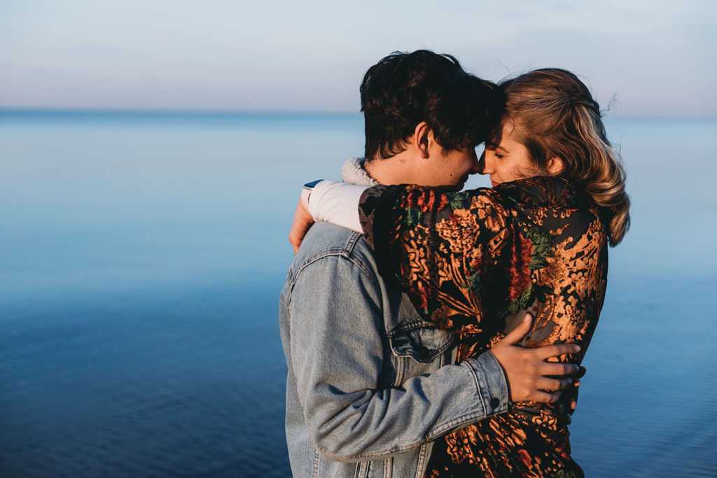 young couple hug in front of still water of Lake Michigan during their Indiana Dunes engagement photos