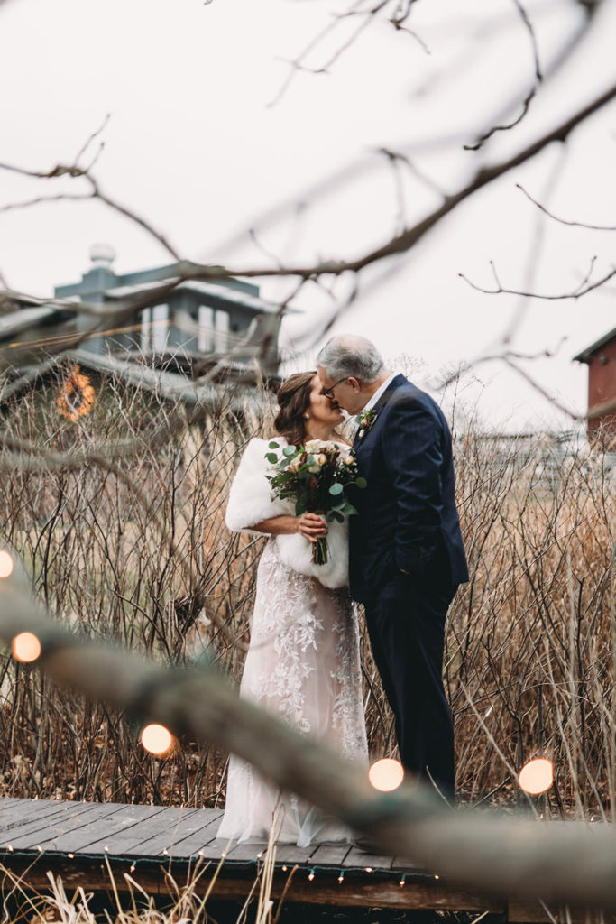 framed by tree branches with fairy lights a bride and groom kiss during their traders point creamery elopement