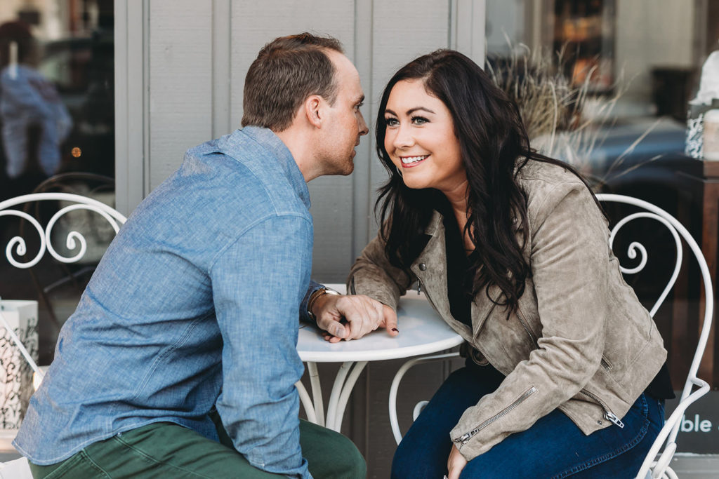 man whispers into woman's ear at little coffee shop table during their downtown zionsville engagement session