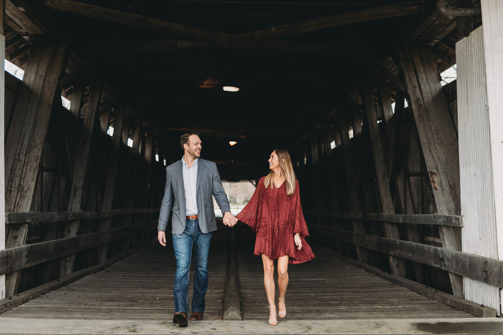 young woman in red dress holds hands with man in suit jacket and jeans on a covered bridge captured by their Noblesville Photographer