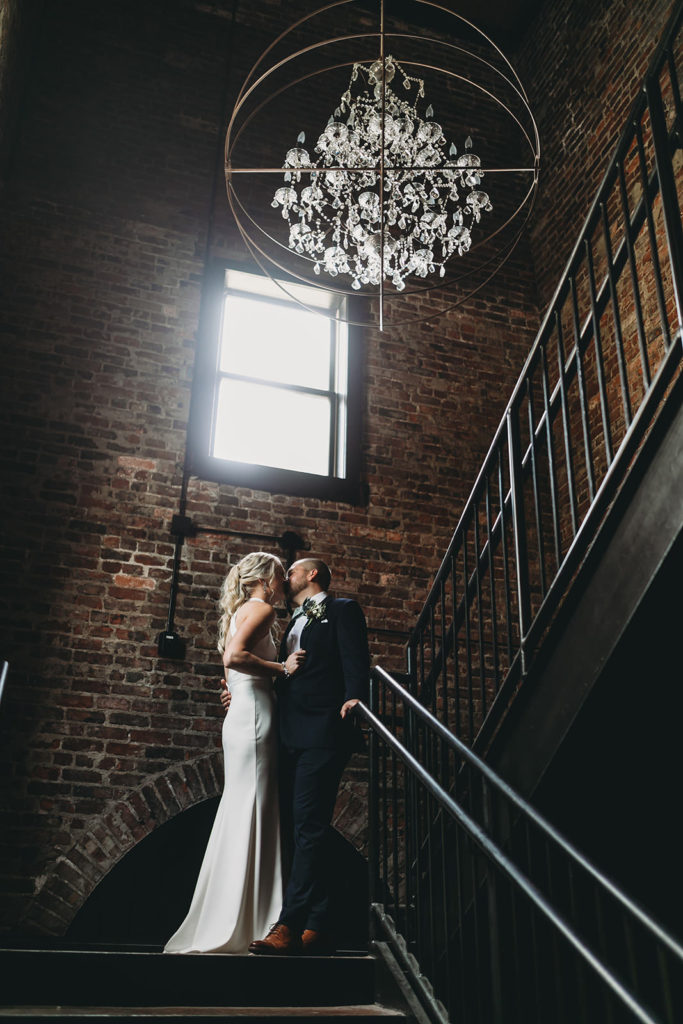 bride and groom kiss under chandelier on stairs at canal 337 before their indy canal wedding