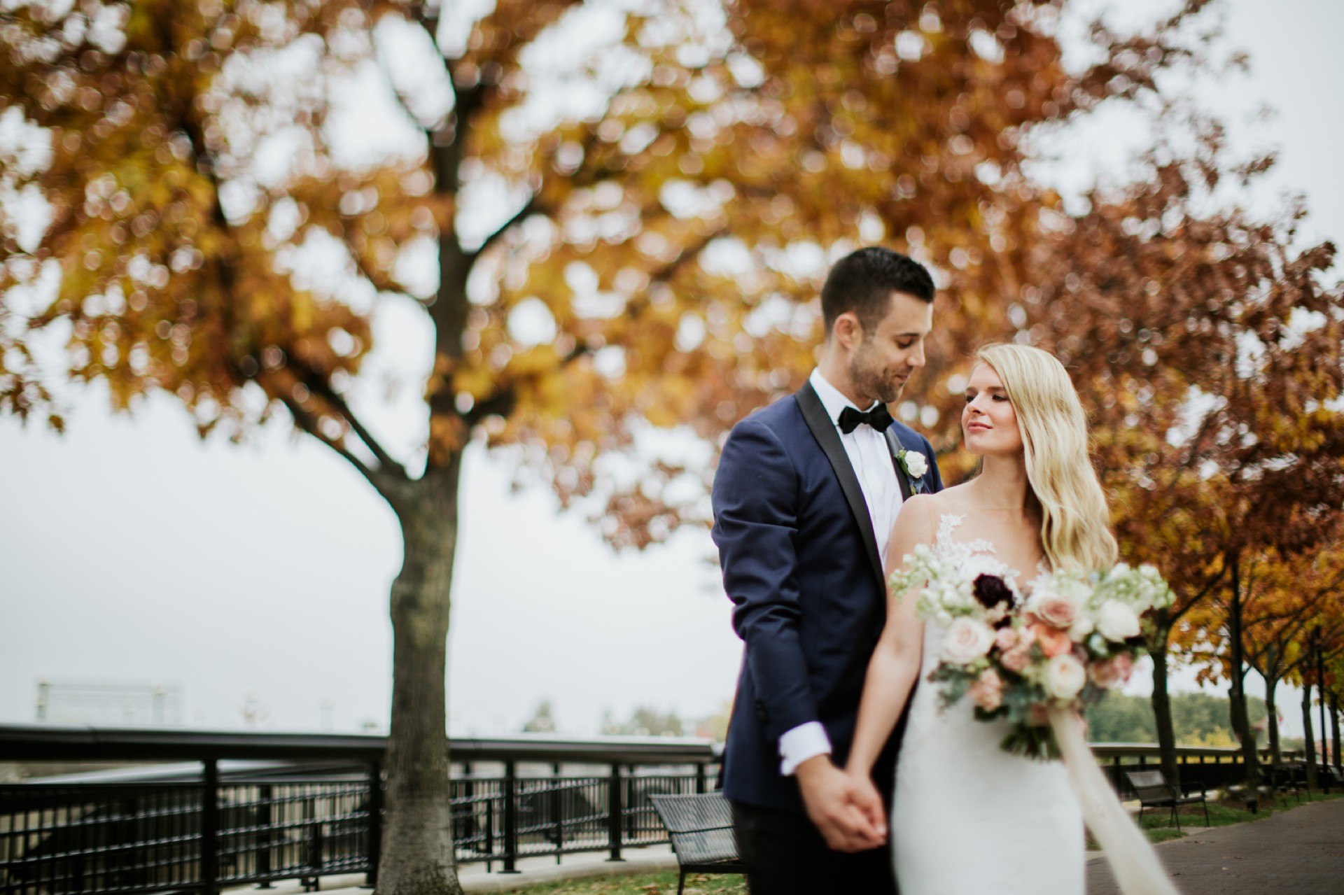 stunning freelensing photo of a bride and groom in the white river park holding hands, the bride's face is in focus but not much else in the photo and leaves on the trees behind them are fading from a brilliant orange to a brown