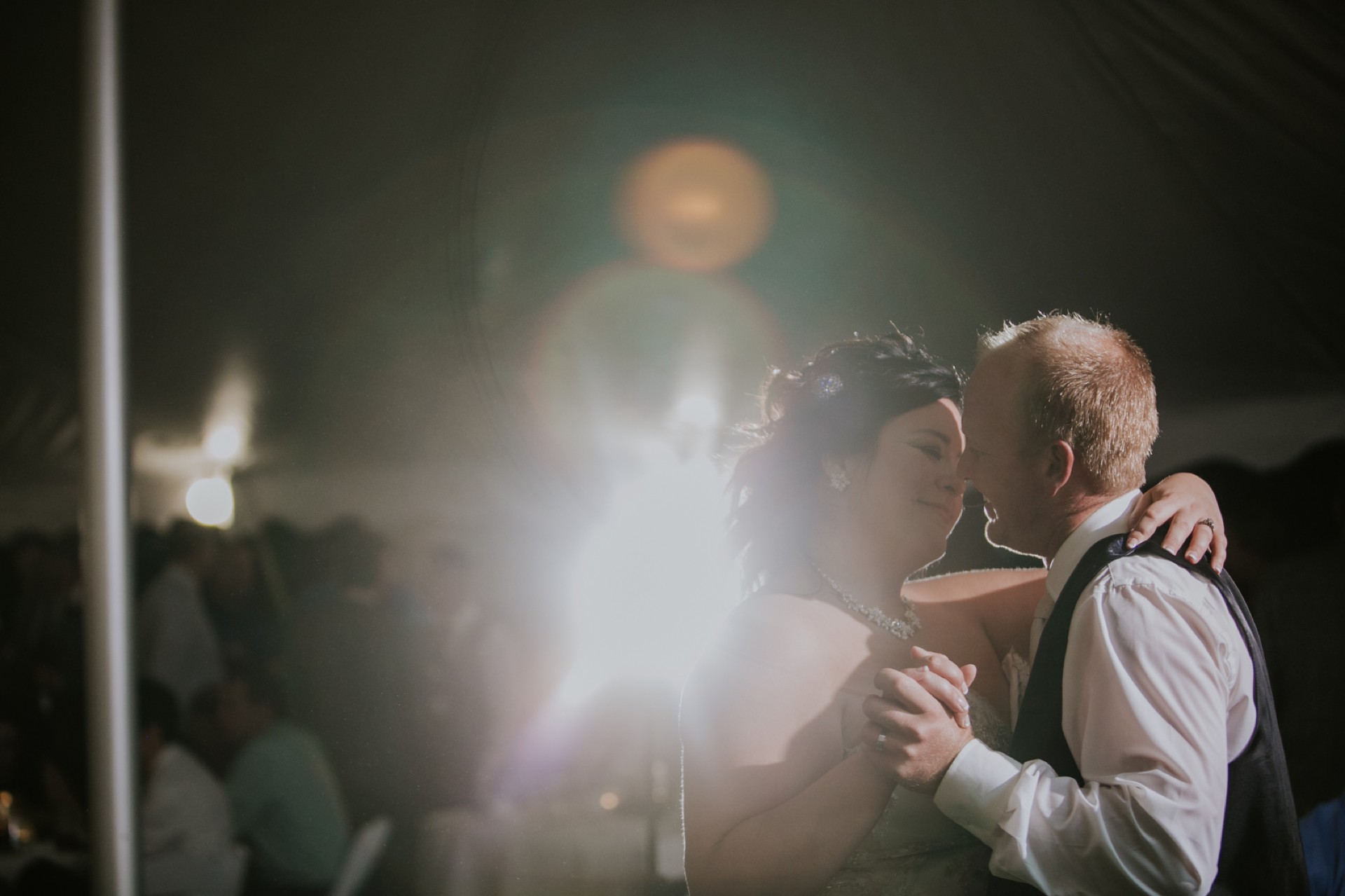 a bride with black hair a bejeweled earrings dances with a red haired man while backlight by off camera flash causing lens flares on the image at their the inn at irwin gardens wedding