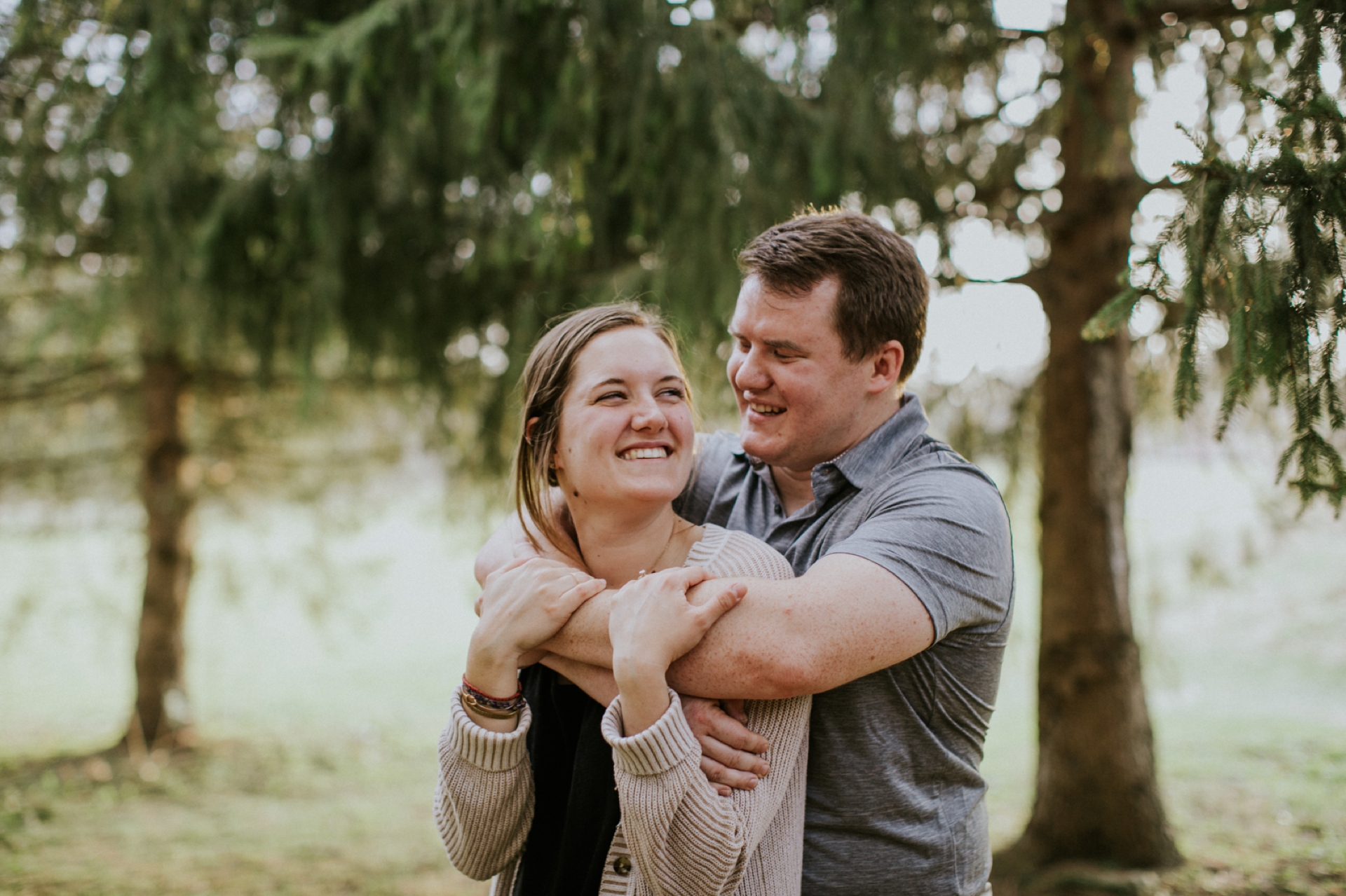man looking at woman under pine trees while hugging her from behind