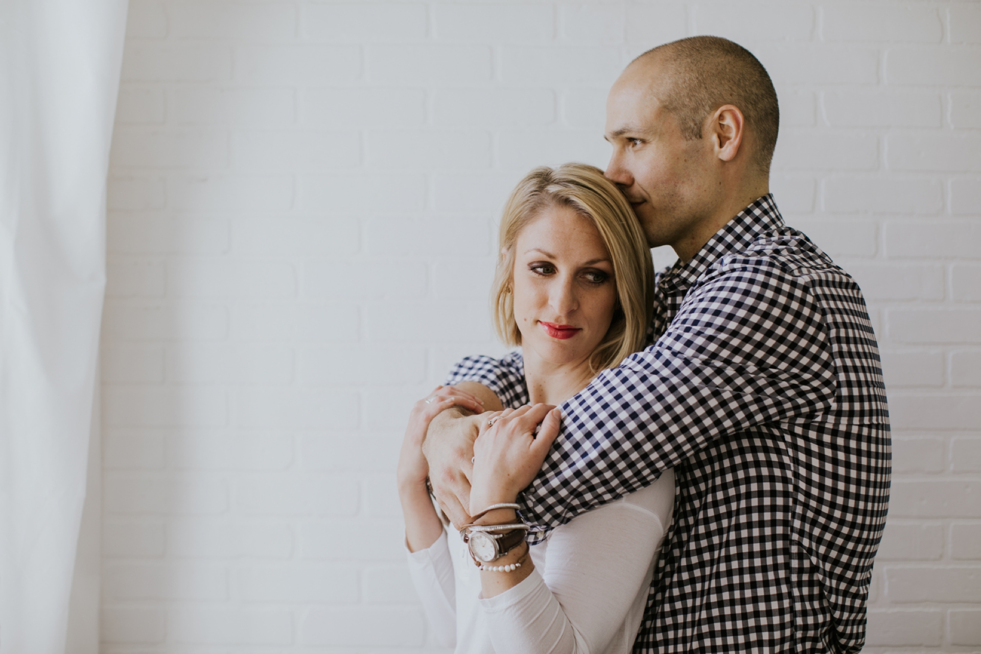 Bald Man Hugs Blonde Woman from Behind in White Room next to Window