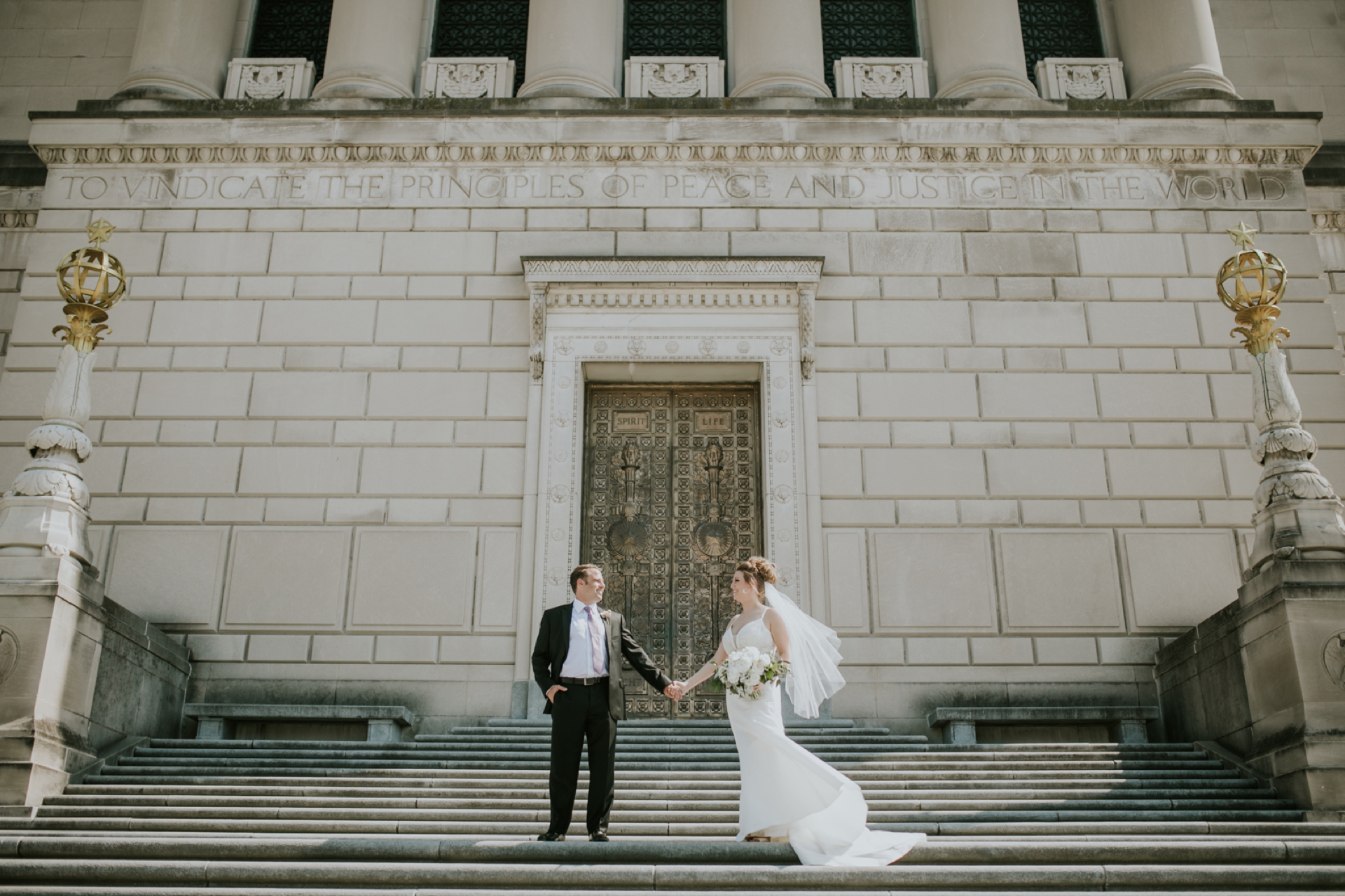 Man in tuxedo and woman in white dress stand on the Indiana War Memorial steps holding hands and looking at each other