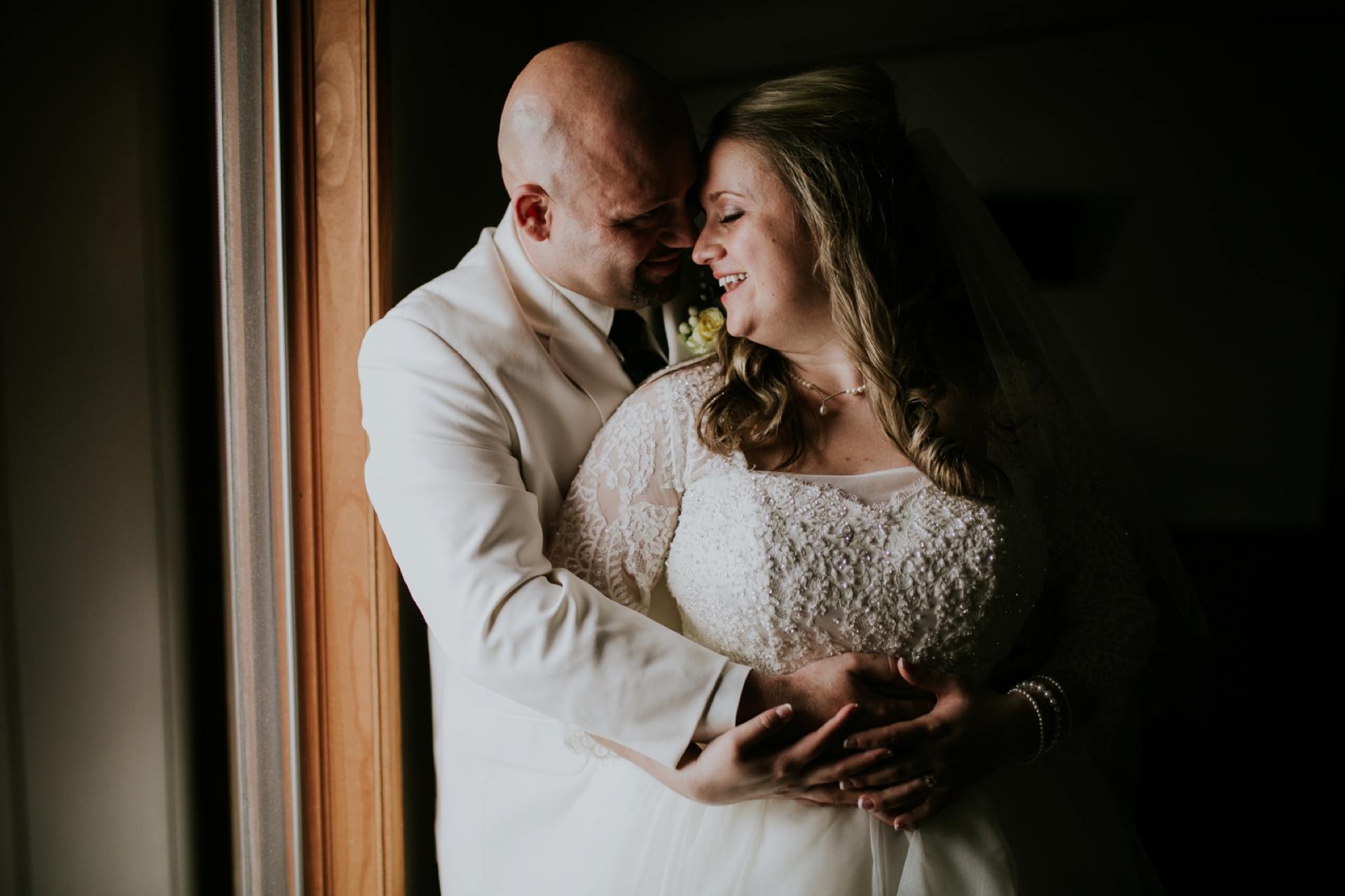 a bride smiles at her groom as he hugs her in moody window light