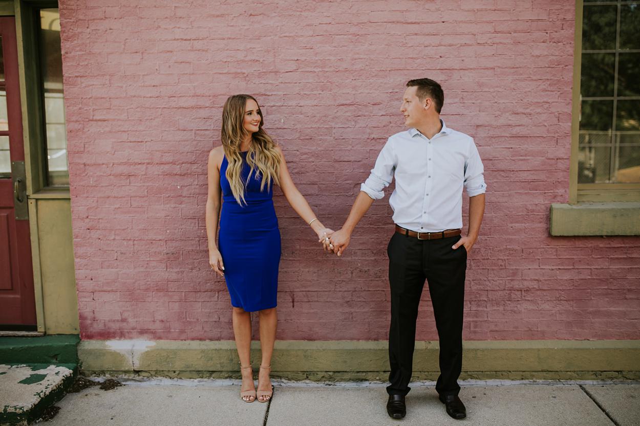 Woman in blue dress and man in slacks hold hands in front of brick wall during their war memorial engagement session