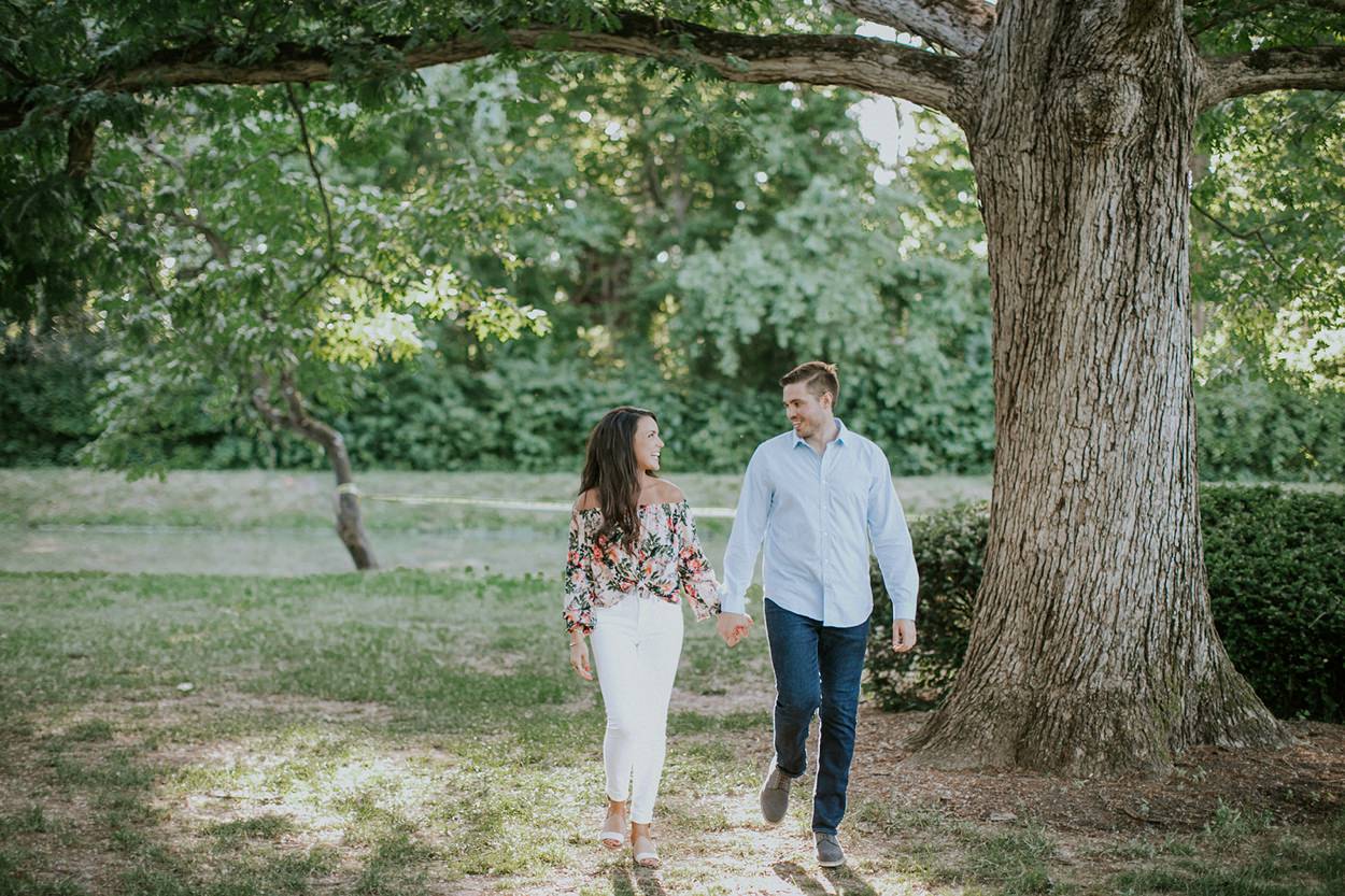 A young couple holds hands while walking under a tree in their holcomb gardens engagement session