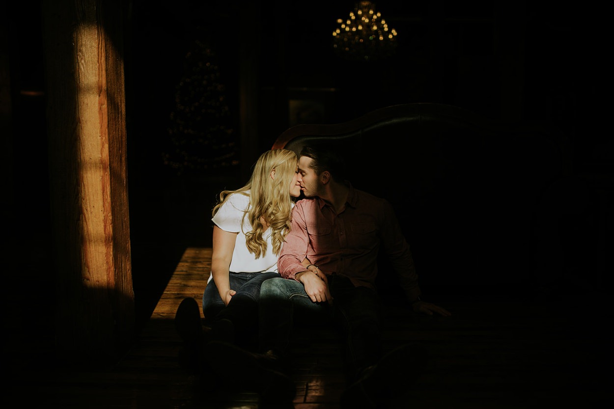 blonde woman sitting in moody window light kisses a man sitting next to her in dark and moody shadow at their mavris engagement session