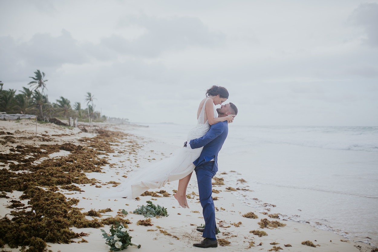 A man in a blue suit lifts a woman in a white dress into the air on a beach at this punta cana destination wedding