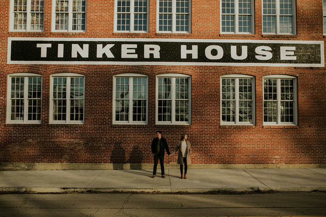 a man and woman stand outside of a brick building for their tinker house engagement session