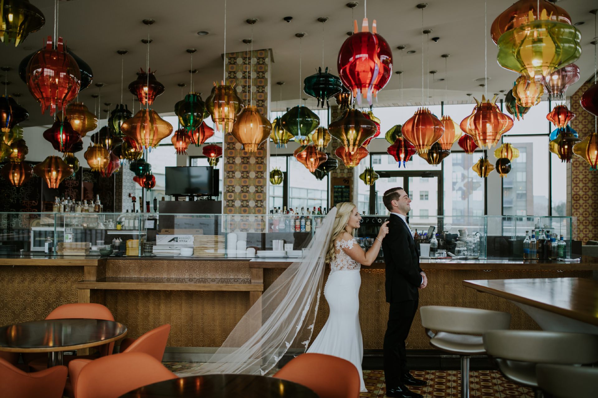 a bride in a white dress taps a groom in a suit on the shoulder under a bunch of hanging glass lamps in a bar in this mavris wedding photography