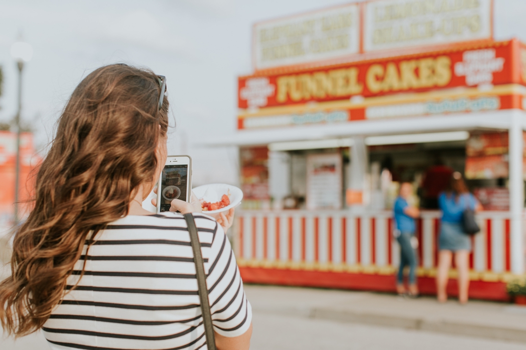 young woman takes photo of funnel cake with phone while funnel cake stand is in her background at the indiana state fair for this fair food photography