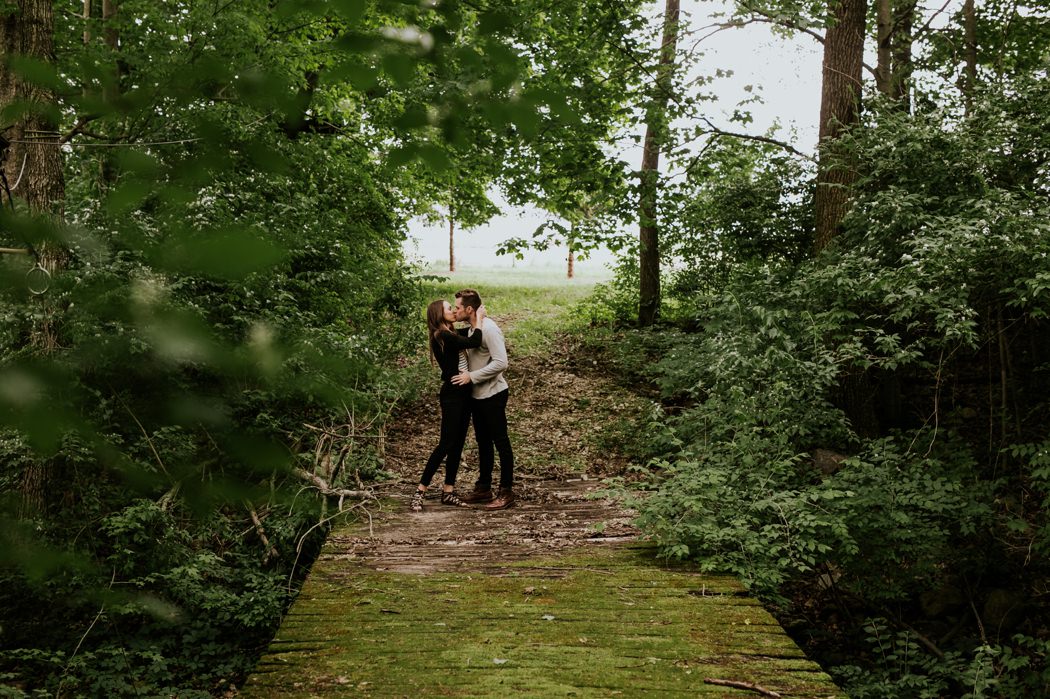 a gorgeous young couple stands at the start of a moss covered wooden bridge in the forest during their zionsville engagement session