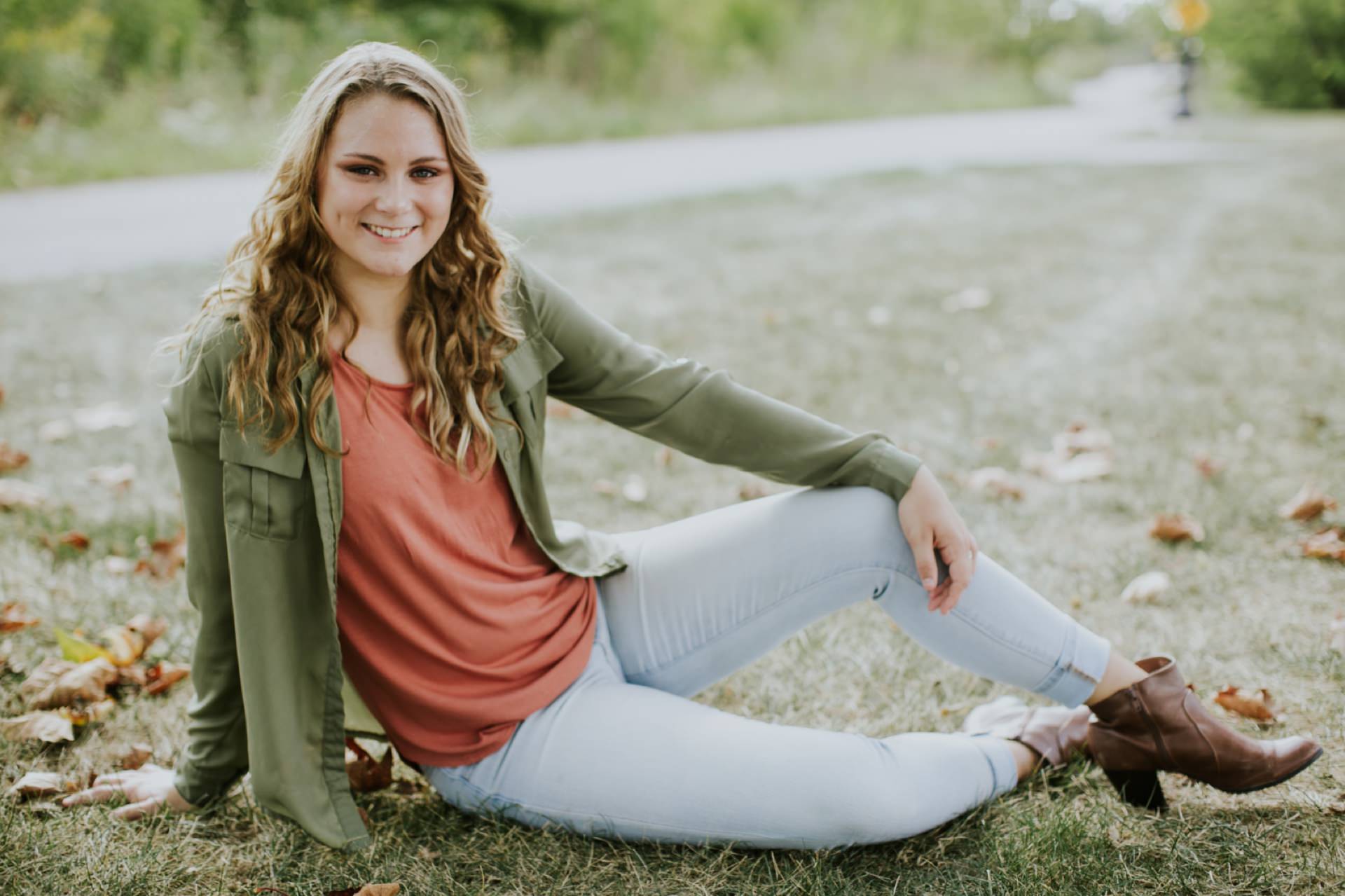 blonde girl in green jacket and jeans sits on grass for Muncie Indiana Senior Portraits
