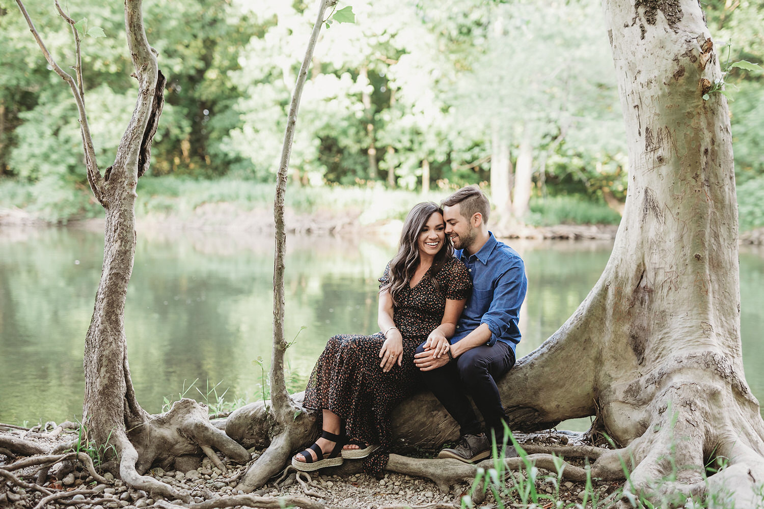 man and woman sit on roots of tree in front of river during their Strawtown Koteewi Park engagement session