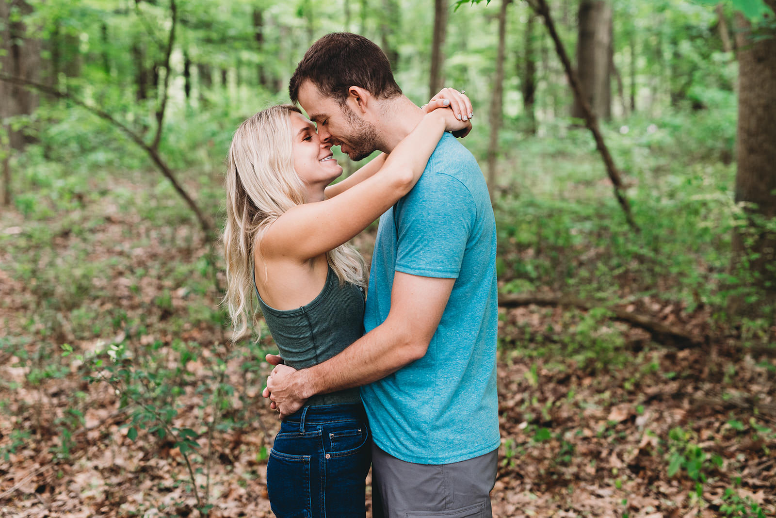 blonde woman in a green tank top and denim jeans kisses a man in shorts with a blue shirt in the woods