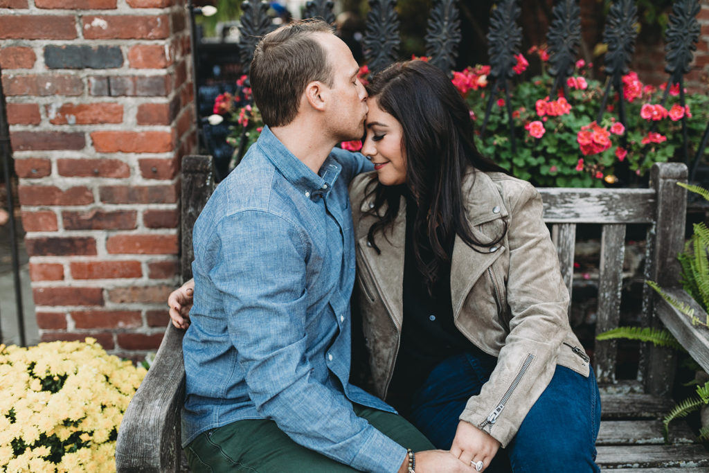 man kisses woman with dark hair in front of red flowers during their downtown zionsville engagement session