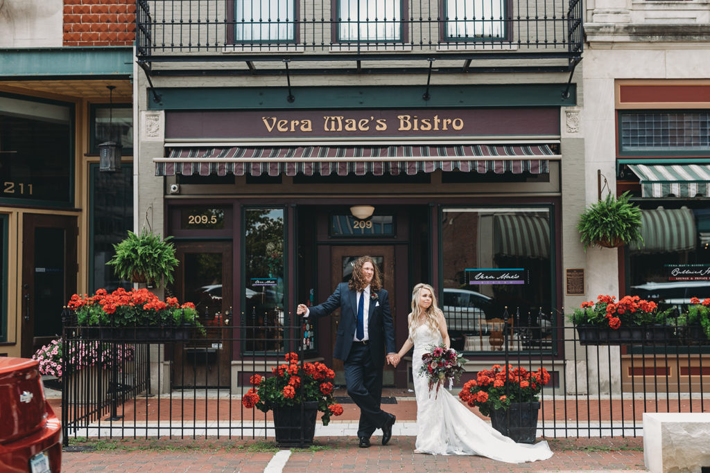 A bride and groom stand outside Vera Mae Bistro in Muncie indiana, along the brick road and black metal fence in their unique wedding photos