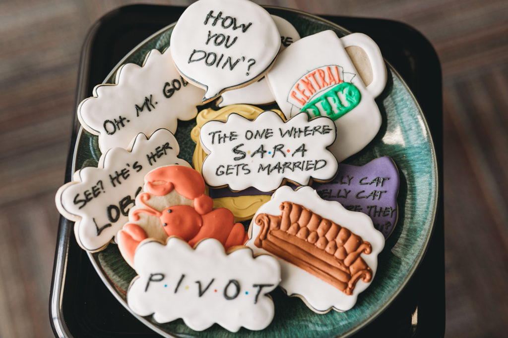 A dozen iced cookies themed as the show Friends, the brides favorite tv show. One has an orange couch, another says "The one where Sara Gets Married", another cookie is a lobster. These are some unique wedding photos