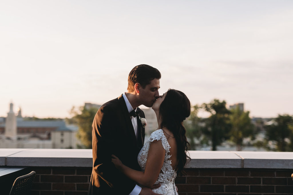 bride and groom kiss on rooftop of embassy suites in south bend with the campus in the background during their Notre Dame wedding