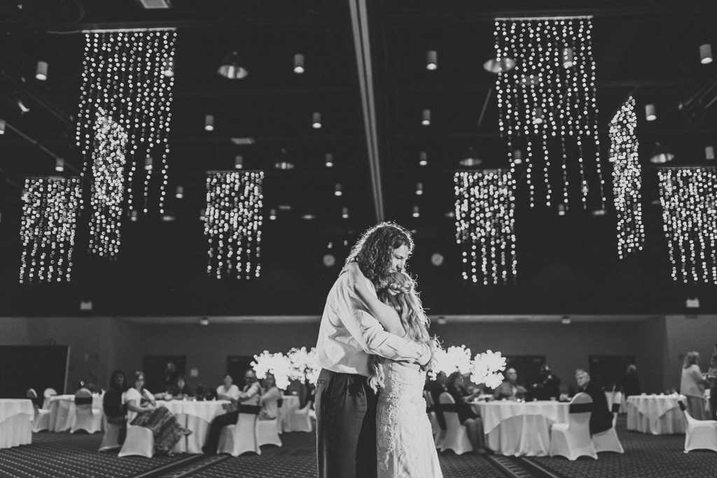 The couple dance in front of their guests. Strings of lights hang from the ceiling at the Horizon Convention Center in their unique wedding photos