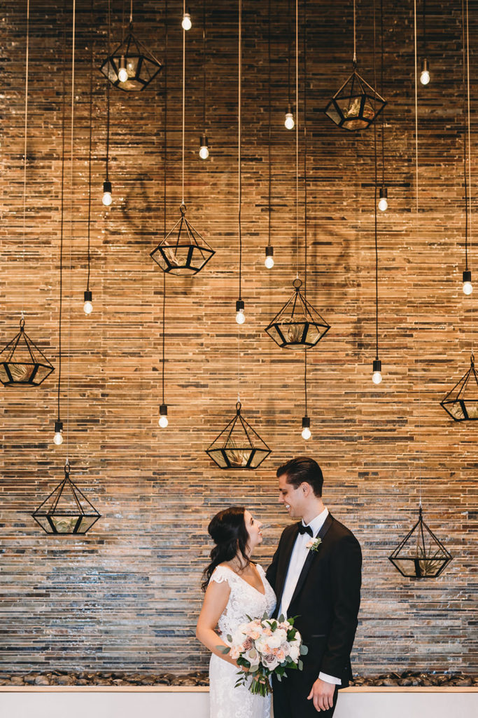 bride and groom in front of mosaic tile wall with hanging plants and hanging lights at embassy suites in south bend before their notre dame wedding