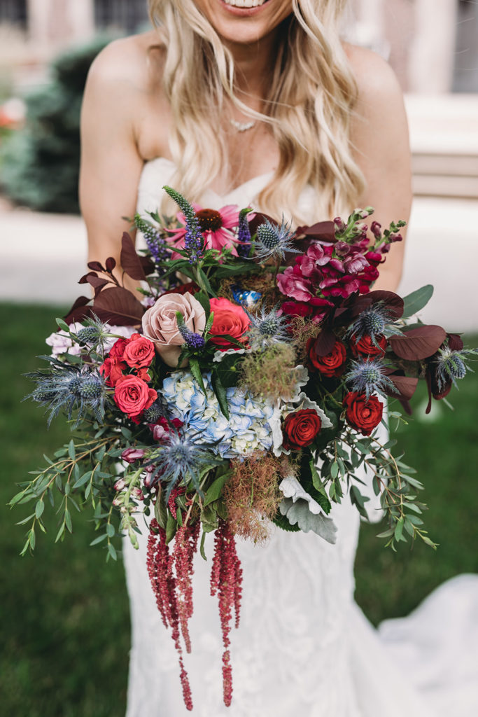An image of the bride holding her flowers where the flowers, red roses, pink roses, blue hydrangeas, other blue and purple flowers and greenery surround outside of the bouquet in her unique wedding photos