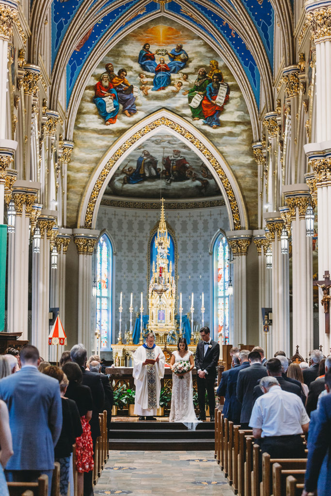wide shot of bride and groom praying with priest at Basilica of the Sacred Heart during their University of Notre Dame wedding