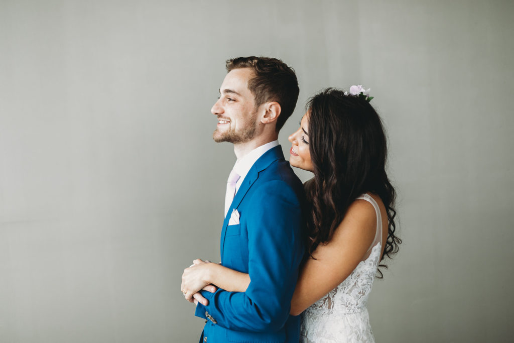 bride hugs groom from behind in front of a white wall at their Indianapolis Hawaiian Jewish Wedding