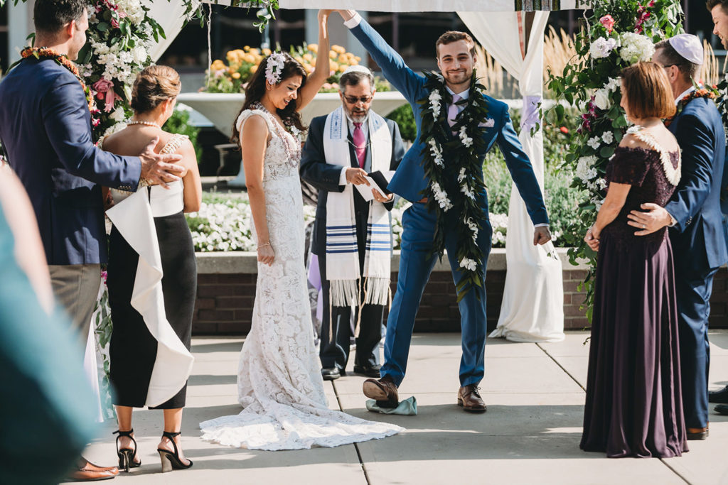 groom holds bride's hand and breaks glass during jewish wedding ceremony while rabbi looks down at the glass at their Indianapolis Hawaiian Jewish Wedding