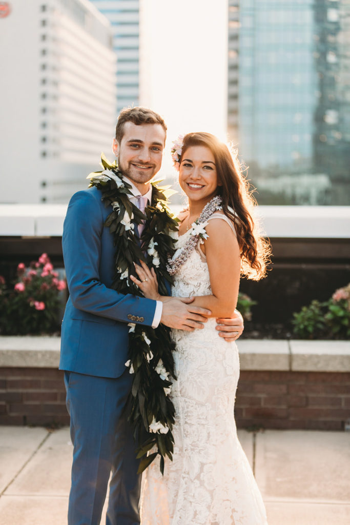 bride and groom both wearing leis smile and hug and look at the camera at sunset on the rooftop of the regions tower during their groom in blue suit dips bride in white dress and kisses her at sunset at their Indianapolis Hawaiian Jewish wedding