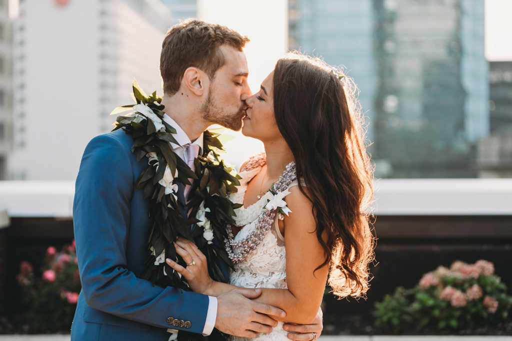 bride and groom in leis backlit by the sunset on a rooftop at the regions tower kiss during their groom in blue suit dips bride in white dress and kisses her at sunset at their Indianapolis Hawaiian Jewish wedding