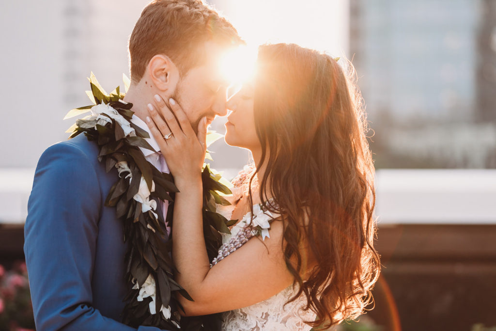 bride and groom touch noses at sunset while wearing leis at their groom in blue suit dips bride in white dress and kisses her at sunset at their Indianapolis Hawaiian Jewish wedding