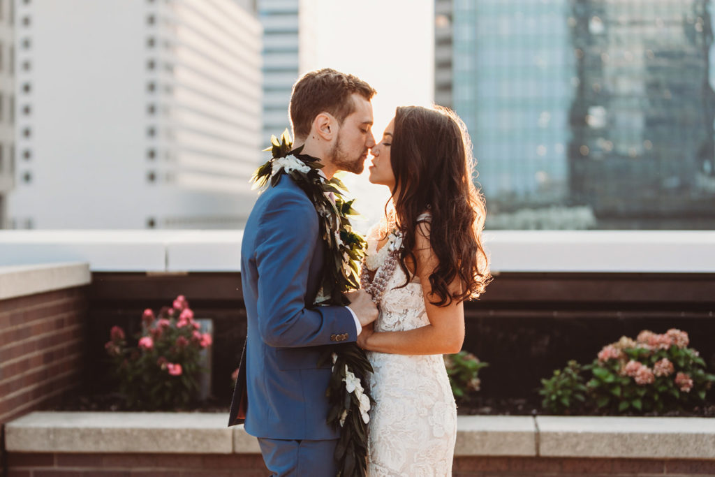 bride and groom wearing leis kiss at sunset at their groom in blue suit dips bride in white dress and kisses her at sunset at their Indianapolis Hawaiian Jewish wedding