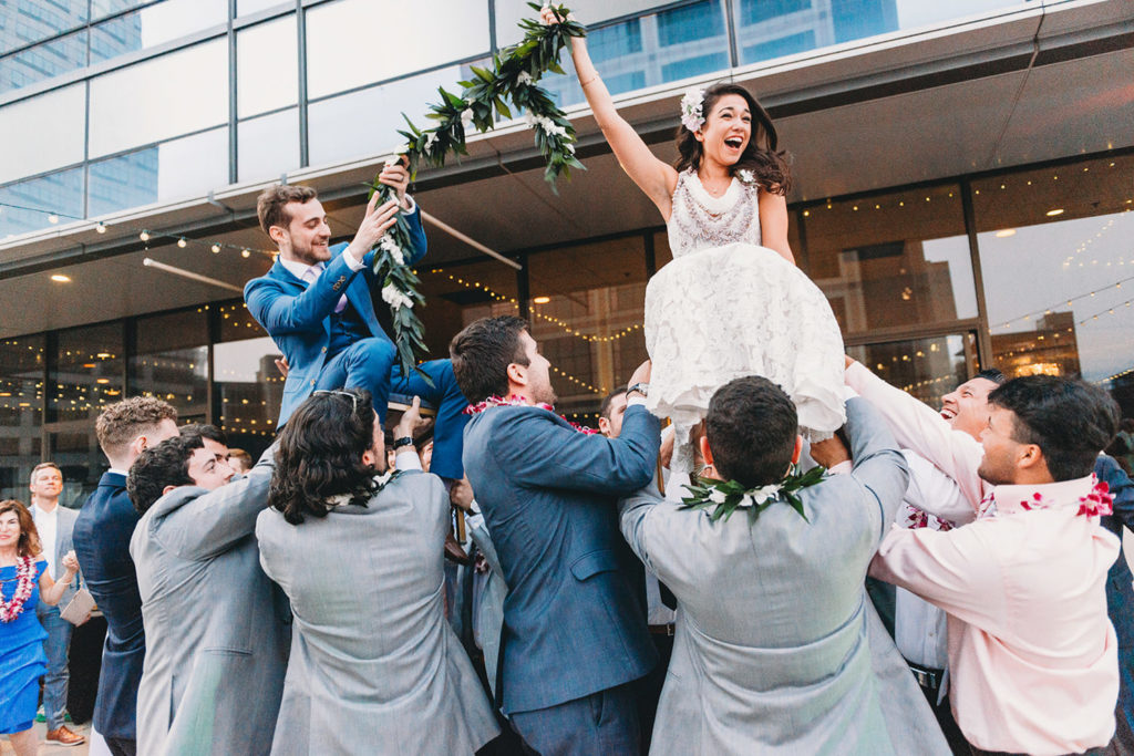 bride and groom laugh and hold lei aloft in air while their guests lift them on chairs at their Indianapolis Hawaiian Jewish Wedding