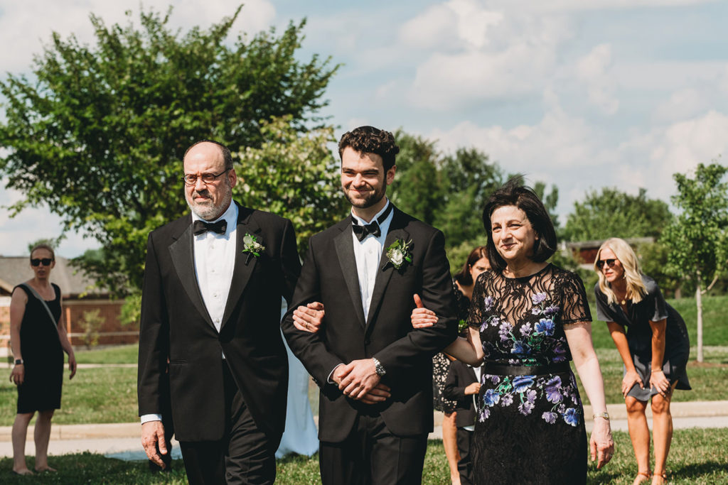 groom walks with parents down aisle to chuppah at his charming coxhall gardens wedding